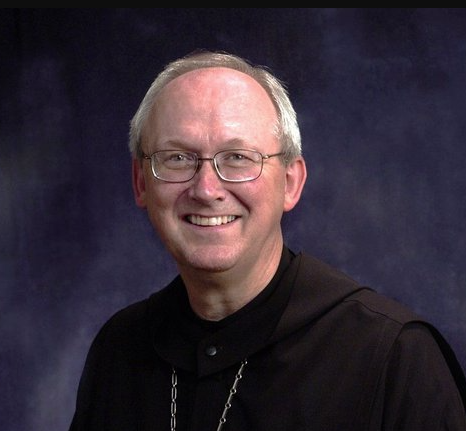 A smiling older man with glasses, short gray hair, and wearing a black robe with a chain necklace, posed in front of a dark blue background.